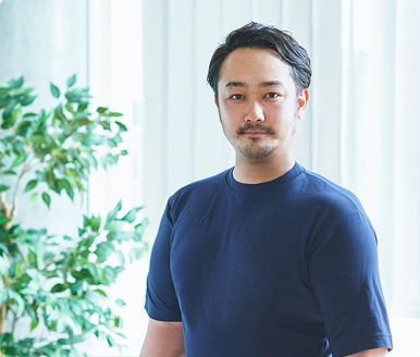 明るい室内で、ネイビーのTシャツを着た男性が落ち着いた表情でまっすぐ立っている。堂々とした佇まいが印象的で、背景には観葉植物とブラインドが見える。